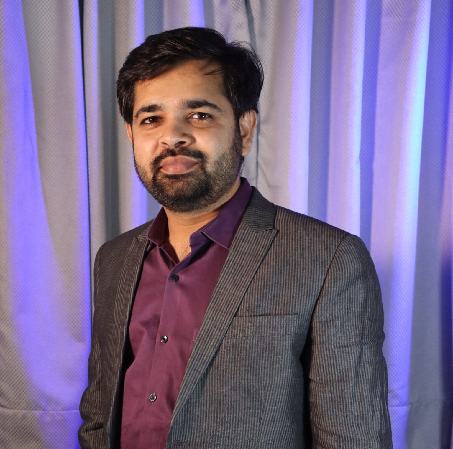 Man in a gray pinstripe blazer and maroon shirt standing indoors with blue-purple stage lighting in the background.