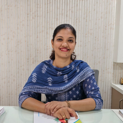 Smiling woman in a blue patterned outfit sits at a desk with papers, hands folded, in an office setting.
