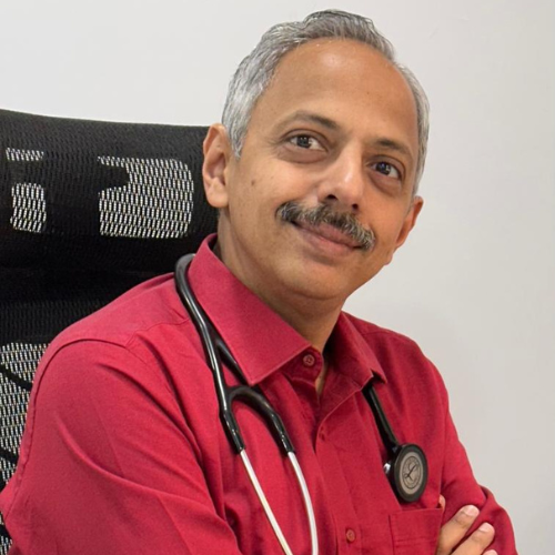 Male doctor with a stethoscope around his neck, wearing a red shirt, seated in a black chair and smiling at the camera.